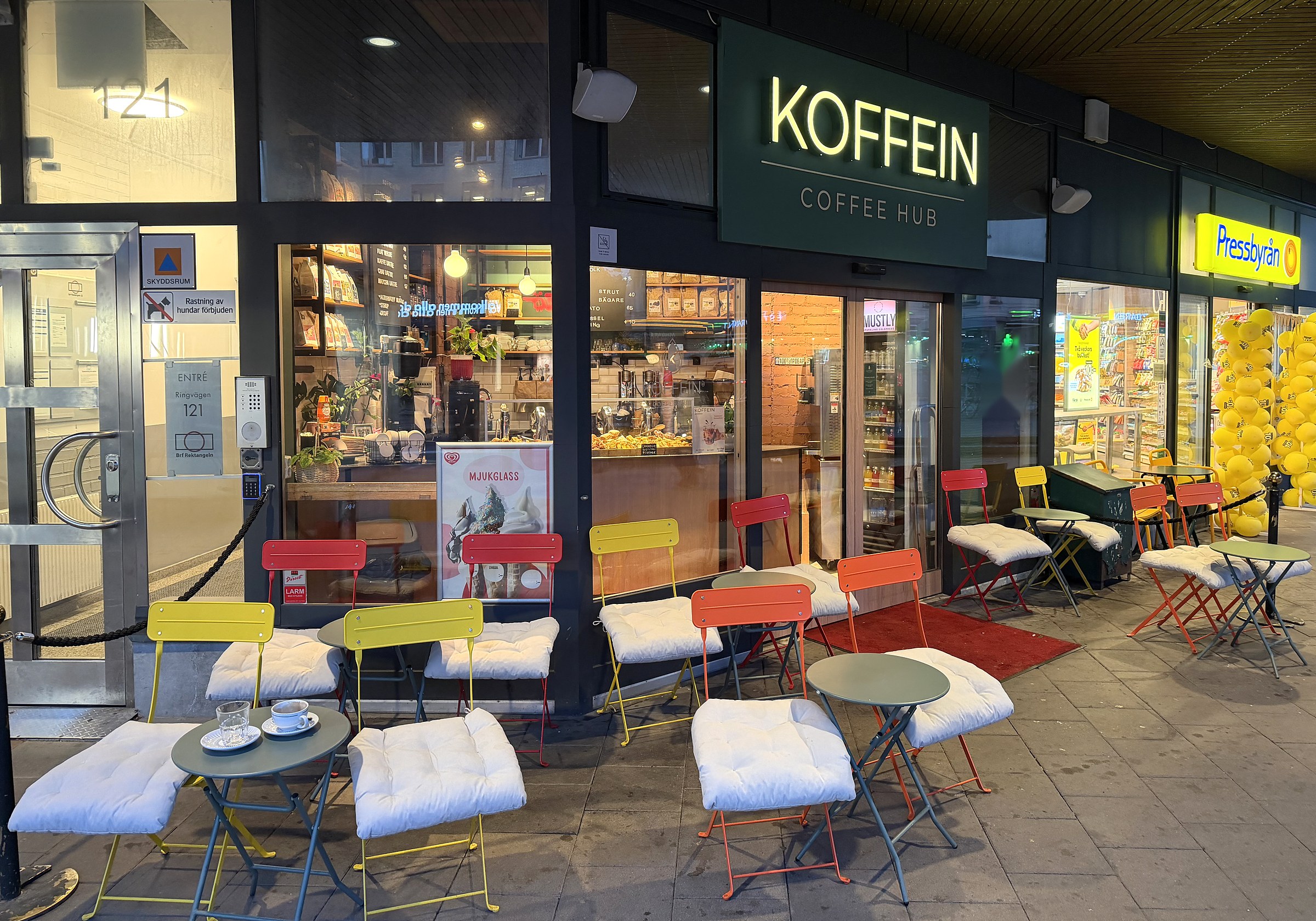 Hela fasaden med Koffeins gröna skylt och alla stolarna ute under arkaden: gula, röda och orange caféstolar med vita dynor och små gröna bord