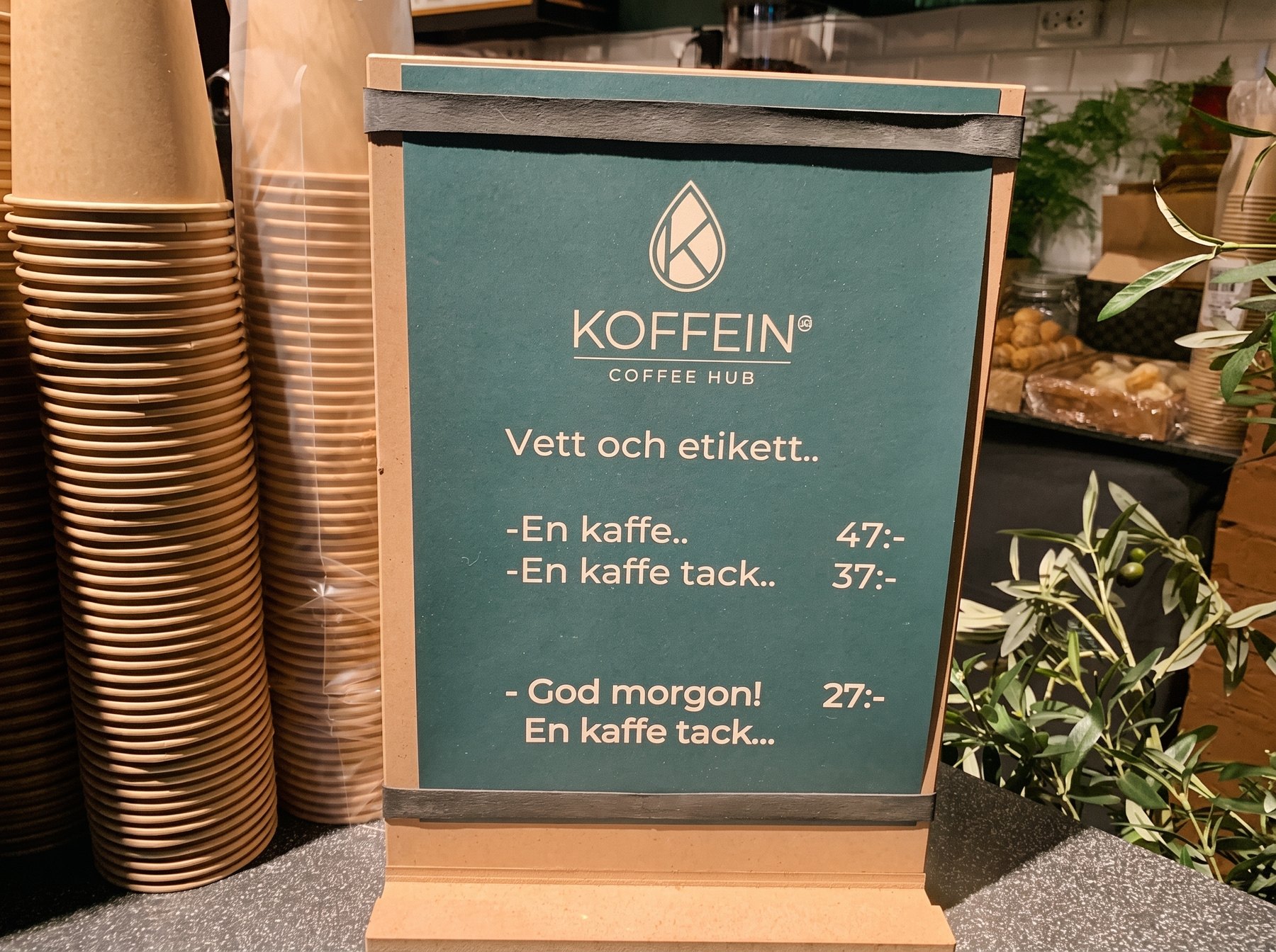 Skylten Vett och etikett vid kassan: En kaffe 47 kr, En kaffe tack 37 kr, God morgon! En kaffe tack 27 kr